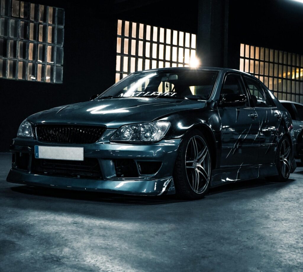A dark colored Lexus IS300 parked in a concrete garage. The lighting exaggerates the aggressive body kit and wing, giving a sporty look.